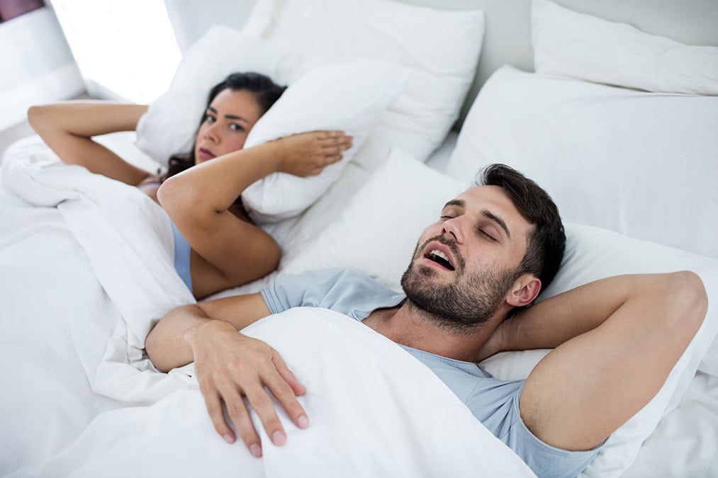 pareja en una cama, mujer se tapa oidos con almohada porque el hombre ronca